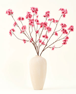 El ramo de flores artificiales de seda Flor de cerezo fucsia de 114 cm añade un vibrante toque de color a tu interior con sus llamativas ramas y flores fucsias, bellamente expuestas en un jarrón de cerámica beige sobre un fondo liso blanquecino.