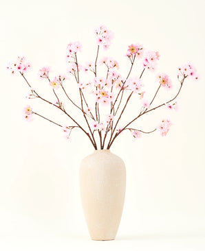 El ramo de flores artificiales de seda Flor de cerezo rosa 114 cm tiene ramas largas y delgadas con racimos de flores de color rosa claro en un jarrón de cerámica beige, que forman un llamativo contraste con el fondo blanco liso.