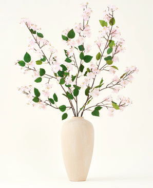 Un jarrón de cerámica beige contiene varias ramas largas con el ramo de flores artificiales Silk de hojas rosas y verdes de Peach Blossom, sobre un fondo blanco liso, perfecto para un interior floral artificial.