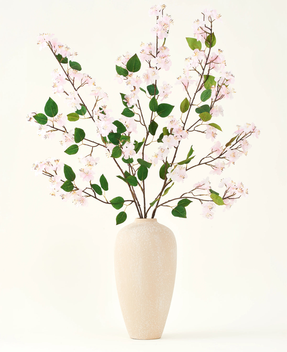 Un jarrón de cerámica beige contiene varias ramas largas con el ramo de flores artificiales Silk de hojas rosas y verdes de Peach Blossom, sobre un fondo blanco liso, perfecto para un interior floral artificial.