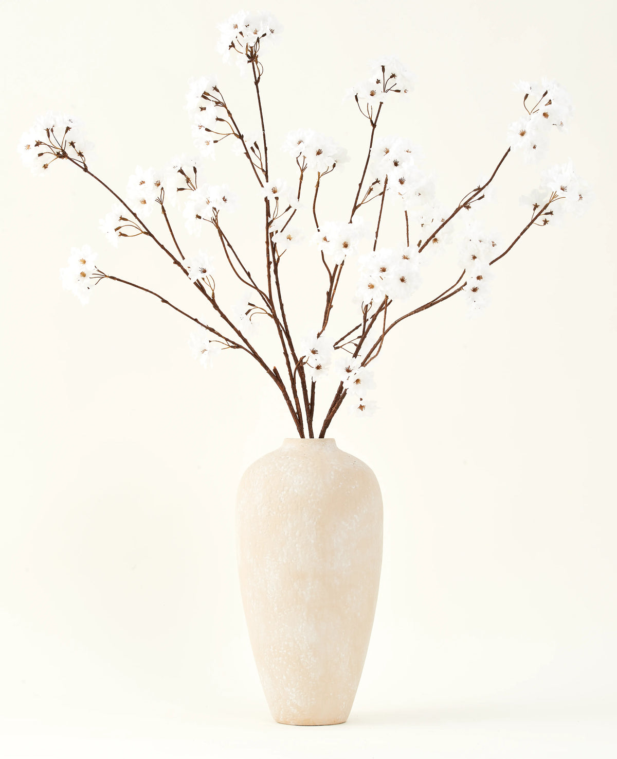 El ramo de flores artificiales de seda Crema de cerezo de 114 cm de altura se encuentra en un jarrón de cerámica beige y aporta una decoración serena a cualquier habitación con sus delicadas ramas de color crema sobre un fondo claro.