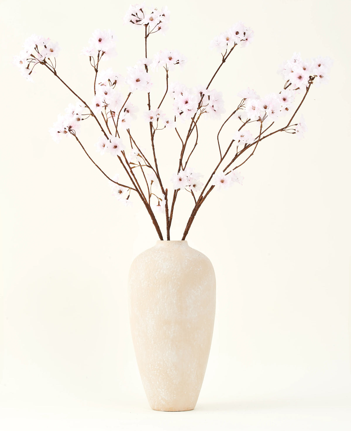 El ramo de flores artificiales de seda Flor de cerezo rosa claro de 114 cm se encuentra en un jarrón alto de cerámica beige con ramas largas y delgadas y delicadas flores de color rosa claro, creando un sereno acento floral artificial sobre un fondo liso.