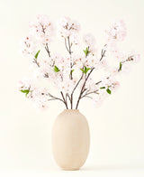 El ramo de flores artificiales de seda Cherry Blossom Blush 119 cm muestra delicadas flores de cerezo de color rosa claro con hojas verdes en un jarrón beige, ideal como decoración floral artificial sobre un fondo claro.