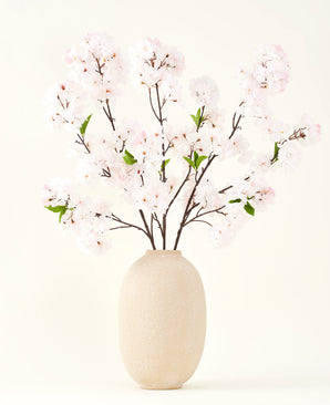 El ramo de flores artificiales de seda Cherry Blossom Blush 119 cm muestra delicadas flores de cerezo de color rosa claro con hojas verdes en un jarrón beige, ideal como decoración floral artificial sobre un fondo claro.