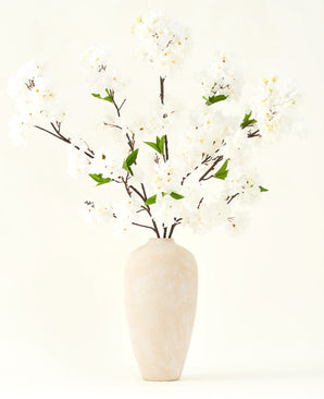 El ramo de flores artificiales de seda Crema de cerezo de 119 cm tiene elegantes ramas marrones con suaves flores artificiales blancas y hojas verdes, expuestas en un sencillo jarrón beige sobre un fondo liso y claro.