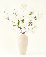 El ramo de flores artificiales de seda Crema de cerezo de 119 cm tiene elegantes ramas marrones con suaves flores artificiales blancas y hojas verdes, expuestas en un sencillo jarrón beige sobre un fondo liso y claro.