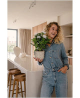 Una mujer sonriente con un mono vaquero se encuentra en una cocina moderna, con la planta artificial blanca Artificial Gardenia UV de 39 cm en sus manos. Los taburetes de madera están alineados a lo largo de una encimera blanca y la luz del sol entra a través de una ventana al fondo.