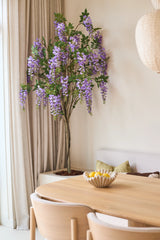 Junto a una ventana con cortinas beige, junto a una mesa de madera con un cuenco de limones y un banco con cojines verdes, se encuentra una planta de glicina artificial de 180 cm con racimos de flores de color azul lila.