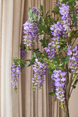 Delante de las cortinas beige cuelga una planta de glicina artificial de 150 cm con hojas verdes.