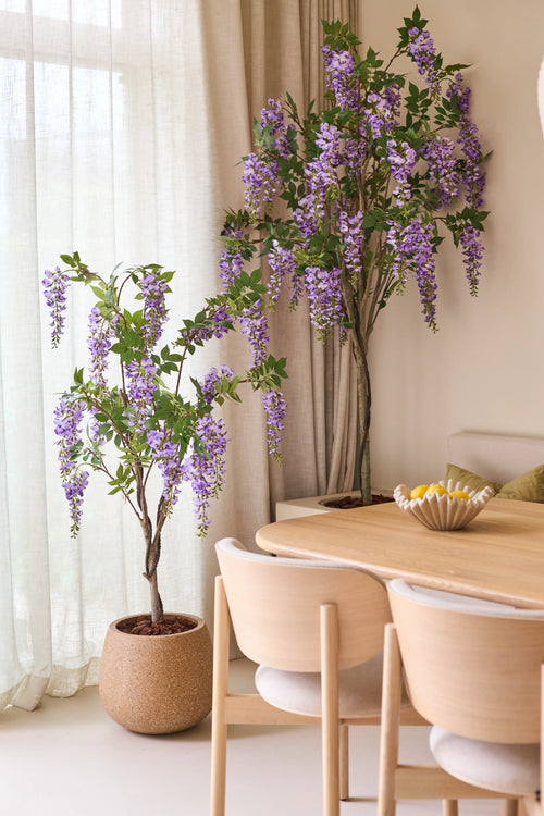 Dos plantas de glicina artificial de 150 cm, con sus flores violetas colgantes, se encuentran junto a una mesa de comedor de madera con sillas claras. La suave luz natural brilla a través de las cortinas translúcidas detrás de las plantas.