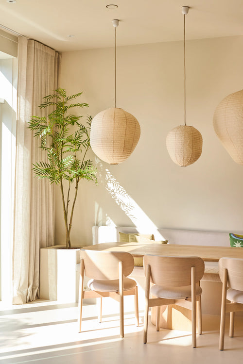 Un comedor luminoso y minimalista está amueblado con sillas de madera clara, una mesa, tres lámparas colgantes de papel, la planta Mahonia artificial de 210 cm en una maceta y cortinas de piso a techo cerca de una gran ventana que deja entrar la luz del sol.