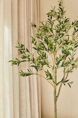 Un olivo artificial de 150 cm, decorado con aceitunas oscuras, queda atractivo en el interior junto a cortinas beige y una pared marrón claro, y proporciona un aspecto tranquilo y natural, ideal para cualquier interior con un olivo.