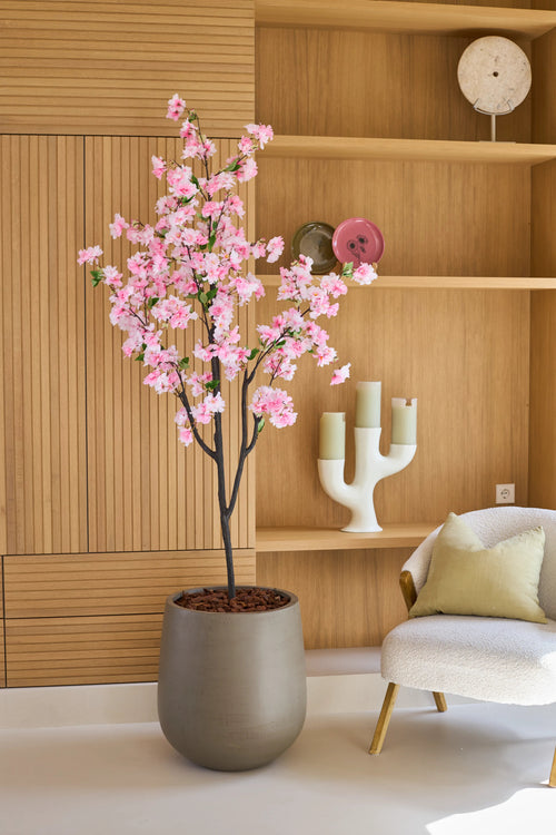 Una flor de cerezo artificial rosa de 180 cm, un árbol en maceta con flores rosas, se encuentra junto a una silla blanca con cojines en una habitación moderna con estantes de madera y objetos decorativos.
