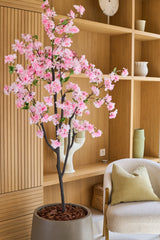 La flor de cerezo artificial rosa de 150 cm, con sus delicadas flores de seda rosa, queda elegante en una habitación moderna con estantes de madera, objetos decorativos, una silla beige y un cojín verde, añadiendo el encanto de las flores de seda a su interior.