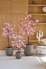 Tres buganvillas artificiales de 180 cm, de color rosa y con flores rosas, se encuentran frente a estantes de madera en la pared, junto a una moderna silla blanca con un cojín.