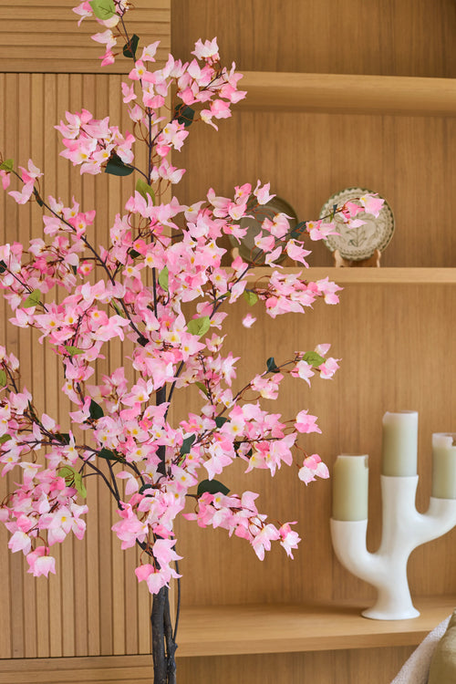 Una planta de buganvilla artificial de 180 cm, de color rosa con flores rosas, se alza ante unos estantes de madera sobre los que se encuentran un plato decorativo y un candelabro blanco con tres velas verdes.