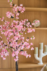 Una planta de buganvilla artificial de 180 cm, de color rosa con flores rosas, se alza ante unos estantes de madera sobre los que se encuentran un plato decorativo y un candelabro blanco con tres velas verdes.