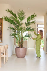 Una persona con un traje verde se encuentra en el interior y toca suavemente las largas hojas de una palmera Kentia artificial de 270 cm, que se encuentra junto a una zona de comedor con sillas de madera clara.