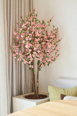 La planta de cerezo artificial rosa de 120 cm con sus flores de cerezo rosas se encuentra junto a una mesa de madera en una habitación luminosa y moderna, con una cortina beige y un sofá claro detrás.