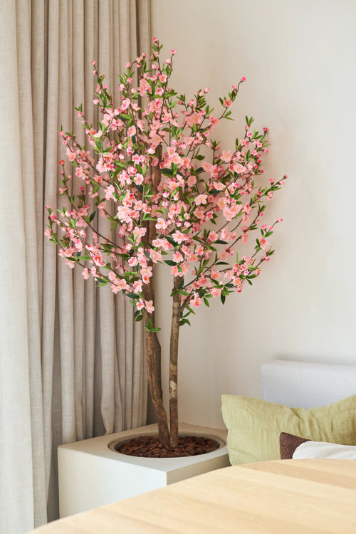 La planta de cerezo artificial rosa de 120 cm con sus flores de cerezo rosas se encuentra junto a una mesa de madera en una habitación luminosa y moderna, con una cortina beige y un sofá claro detrás.
