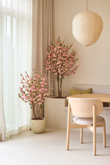Una habitación minimalista con muebles de madera clara, cortinas en color crema y dos Flores de Cerezo Artificiales de 150 cm en color rosa, que aportan flores rosas. Una linterna de papel redonda cuelga sobre la mesa del comedor, con una silla acolchada y un banco incorporado.