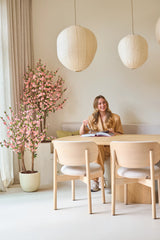 Una mujer está sentada sonriendo ante una mesa con un libro abierto. La habitación está decorada con muebles de madera clara, faroles de papel, una suave luz natural que entra a través de cortinas altas y una planta de cerezo artificial rosa de 120 cm que añade un toque de flor rosa.