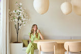 Una mujer con un traje verde claro está sentada en un banco junto a una mesa de comedor de madera, con una taza en la mano. Sobre ella cuelgan farolillos de papel decorativos y en la esquina se alza la flor de cerezo artificial blanca de 180 cm, mientras que una suave luz natural entra a raudales a través de una ventana.