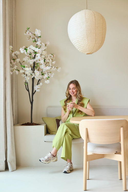 Una mujer con un traje verde claro está sentada en un banco, sonriendo y sosteniendo una taza en la mano. Junto a ella está la Flor de Cerezo Artificial de 180 cm blanca, colocada en una habitación moderna y minimalista con tonos neutros y una linterna de papel redonda sobre su cabeza.