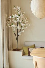 Una flor de cerezo artificial de 180 cm en color blanco se encuentra junto a una zona de estar con cojines en tonos verdes y marrones, cerca de una mesa redonda de madera y una linterna de papel en una habitación suavemente iluminada con tonos neutros.