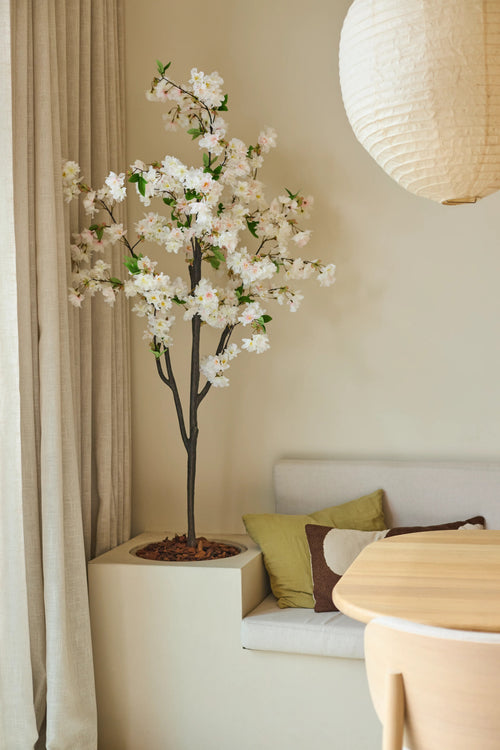 Una flor de cerezo artificial de 180 cm en color blanco se encuentra junto a una zona de estar con cojines en tonos verdes y marrones, cerca de una mesa redonda de madera y una linterna de papel en una habitación suavemente iluminada con tonos neutros.