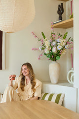 Una mujer con un traje color crema está sentada en un banco con una taza en la mano y una sonrisa en el rostro. Detrás de ella se encuentra un gran jarrón con el ramo de flores artificiales de seda 'Lots of Love XL', compuesto por flores rosas, blancas y amarillas, y al fondo estantes en los que se exponen objetos decorativos. El entorno parece acogedor y moderno.