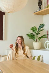 Una mujer sonriente con cabello largo y rubio sentada en una habitación moderna y luminosa con una taza rosa en la mano. A su alrededor hay plantas verdes, incluida la exuberante planta Fénix artificial de 85 cm, así como estantes de madera y objetos decorativos.