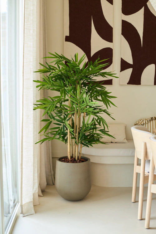 Una planta de bambú artificial de 120 cm se encuentra en un salón moderno y luminoso, cerca de una ventana con visillos, junto a una silla blanca y un arte abstracto en la pared.