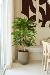 Una planta de bambú artificial de 120 cm se encuentra en un salón moderno y luminoso, cerca de una ventana con visillos, junto a una silla blanca y un arte abstracto en la pared.