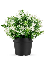 La Planta Artificial Trébol de Cuatro Hojas blanco UV 22 cm es una maceta negra con hojas verdes artificiales y pequeñas flores blancas sobre un fondo liso blanco.
