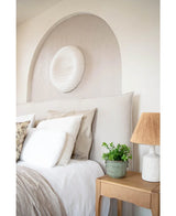 Un dormitorio acogedor con una cabecera beige, ropa de cama y almohadas neutras, una mesita de noche de madera cubierta con una planta de eucalipto artificial de 27 cm, una lámpara con una pantalla texturizada y un arte redondo en la pared blanca sobre la cama en un nicho abovedado.