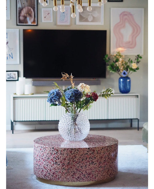 Un salón moderno tiene una mesa de centro de terrazo con un jarrón de cristal con flores, un televisor en la pared, cuadros y fotografías enmarcadas, un aparador blanco y la Hortensia Artificial Deluxe Lila de 57 cm como elegante arreglo floral azul al fondo.