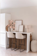 Un bar hogareño minimalista con dos taburetes beige con cojines, un mostrador blanco, arte enmarcado, una lámpara de mesa, decoración en tonos neutros y flor de cerezo artificial de 100 cm de color rosa claro en jarrones contra una pared de color claro.
