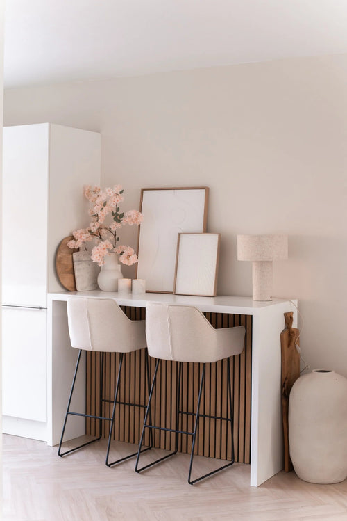 Un bar hogareño minimalista con dos taburetes beige con cojines, un mostrador blanco, arte enmarcado, una lámpara de mesa, decoración en tonos neutros y flor de cerezo artificial de 100 cm de color rosa claro en jarrones contra una pared de color claro.
