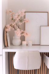 Un escritorio minimalista con una silla blanca, dos velas, bandejas decorativas, arte abstracto enmarcado y una flor de cerezo artificial de 100 cm de color rosa claro en un jarrón blanco sobre una superficie blanca.