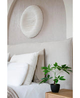 Una cama cuidadosamente hecha con ropa de cama blanca se encuentra junto a una mesita de noche de madera con la planta Pachira Artificial de 30 cm. Encima de la cama, un cuadro circular estructurado en blanco decora la pared de color claro.
