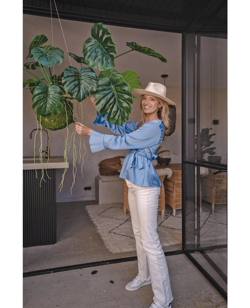 En el interior hay una mujer sonriente con sombrero, blusa azul y pantalón blanco que sostiene una planta artificial colgante Monstera Deluxe de 60 cm con grandes hojas verdes, rodeada de una decoración moderna para un ambiente tropical con plantas decorativas.