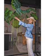 Una mujer sonriente con blusa azul, pantalones blancos y sombrero muestra la planta colgante artificial Monstera Deluxe 80 cm con enredaderas verdes sinuosas, que aporta una atmósfera tropical al luminoso espacio interior con paredes de cristal y muebles de ratán.