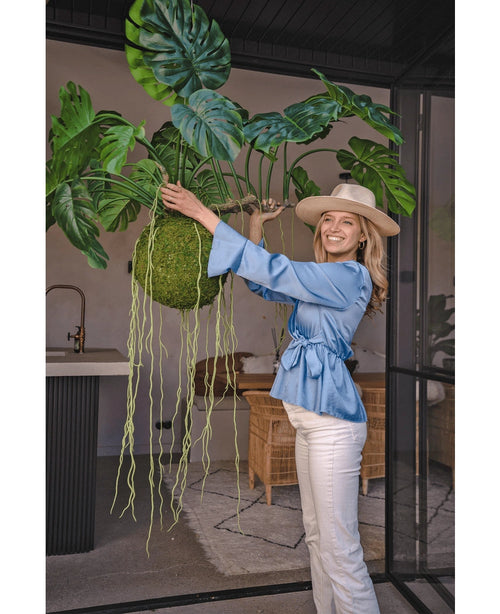 Una mujer sonriente con blusa azul, pantalones blancos y sombrero muestra la planta colgante artificial Monstera Deluxe 80 cm con enredaderas verdes sinuosas, que aporta una atmósfera tropical al luminoso espacio interior con paredes de cristal y muebles de ratán.