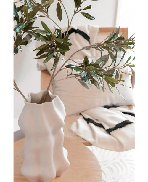 Un jarrón blanco esculpido sostiene una hoja de olivo Art de 90 cm UV con hojas verdes sobre una mesa de madera clara. Detrás, una silla beige y dos cojines de color crema con llamativas rayas negras realzan el ambiente mediterráneo.