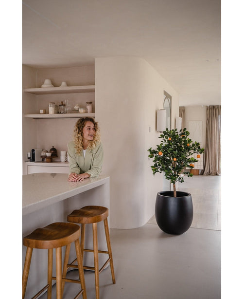 Una mujer de pelo rubio rizado sonríe detrás de un mostrador con dos taburetes de madera. El Naranjo artificial de 120 cm en maceta aporta encanto a un espacio moderno y minimalista decorado en tonos neutros.