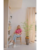 Una mujer con un suéter rosa y jeans sentada en una silla de madera junto a una planta de helecho artificial de 120 cm, con estanterías y una ventana con cortinas beige en una habitación luminosa y moderna.