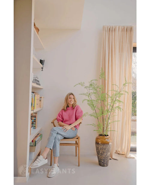 Una mujer con un suéter rosa y jeans sentada en una silla de madera junto a una planta de helecho artificial de 120 cm, con estanterías y una ventana con cortinas beige en una habitación luminosa y moderna.