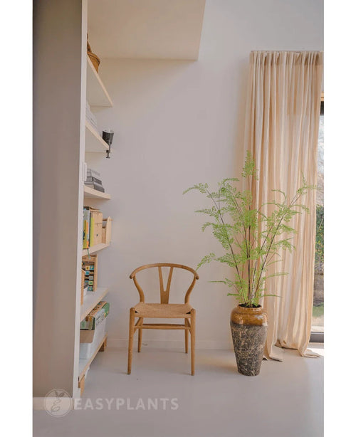 Junto a un helecho artificial de 120 cm de altura en una maceta hay una silla de madera, cerca de cortinas color crema y una pared con estanterías llenas de libros y adornos. La luz del sol ilumina la habitación minimalista, donde elegantes plantas de interior como Art Fern añaden un toque de tranquilidad inspirada en la naturaleza.
