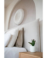 Un dormitorio moderno tiene una cabecera tapizada en color beige, almohadas blancas, una suculenta artificial de 20 cm sobre una mesita de noche de madera y un arte de pared con textura redonda sobre la cama.